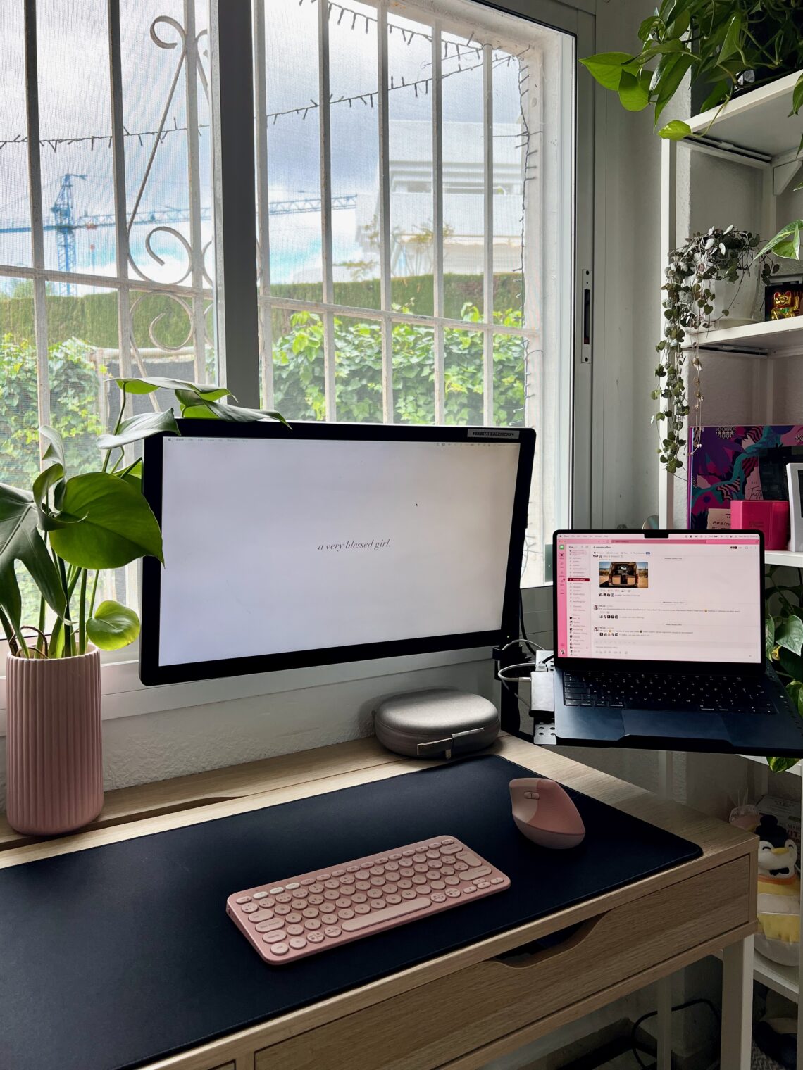Escritorio minimalista de oficina en casa con monitor elevado, laptop, soporte ergonómico, teclado inalámbrico y plantas decorativas junto a una ventana