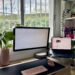 Escritorio minimalista de oficina en casa con monitor elevado, laptop, soporte ergonómico, teclado inalámbrico y plantas decorativas junto a una ventana