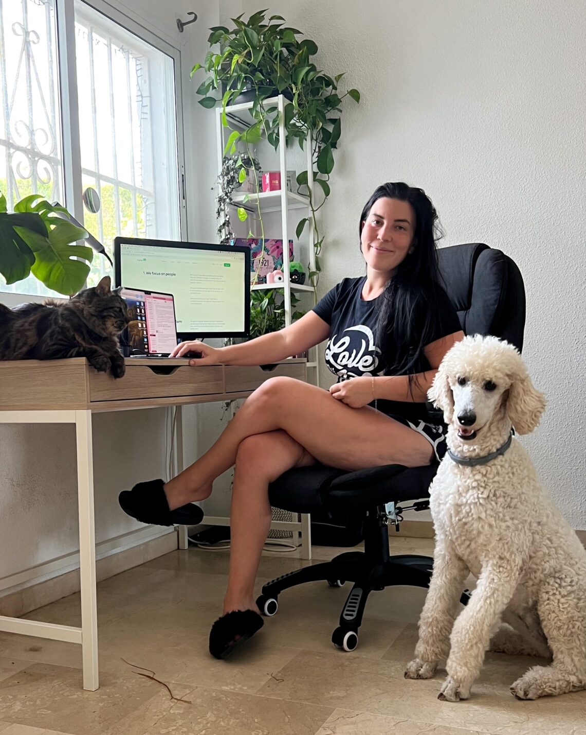 Woman working remotely at her desk with a cat on the table and a dog beside her, surrounded by plants