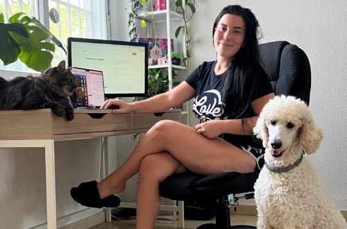Woman working remotely at her desk with a cat on the table and a dog beside her, surrounded by plants