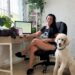 Woman working remotely at her desk with a cat on the table and a dog beside her, surrounded by plants